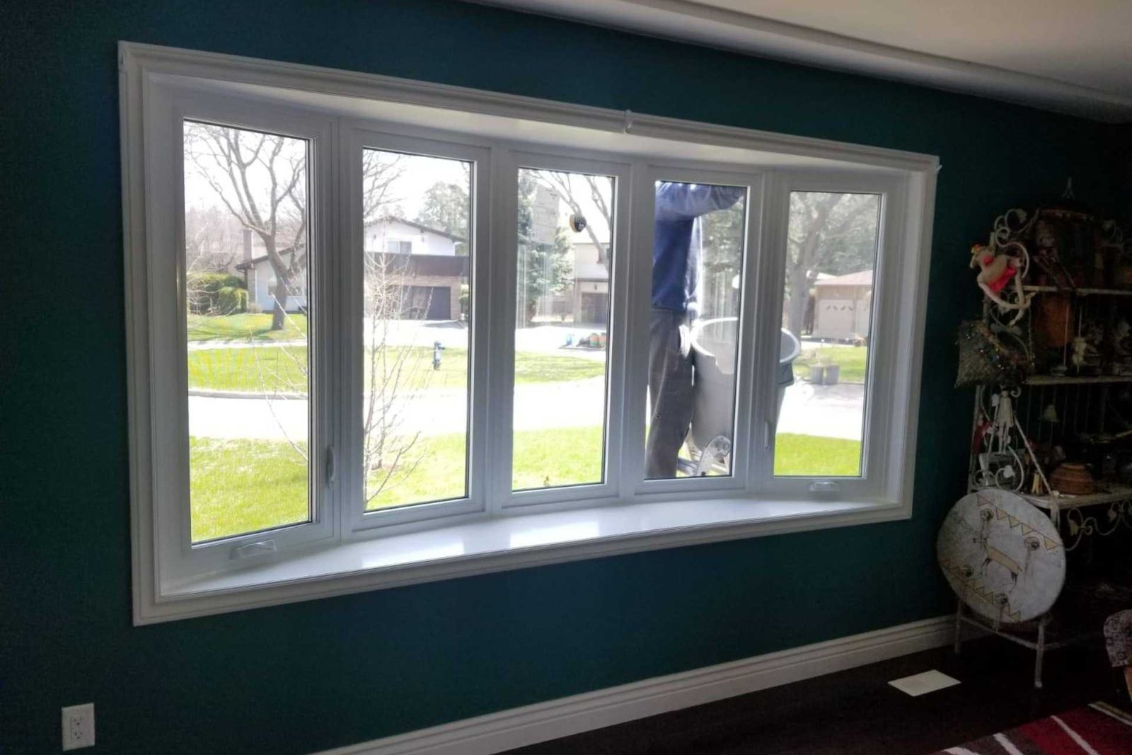 Interior view of a white-framed curved bay window with multiple glass panels, showing a person cleaning the exterior glass and a green neighborhood outside.