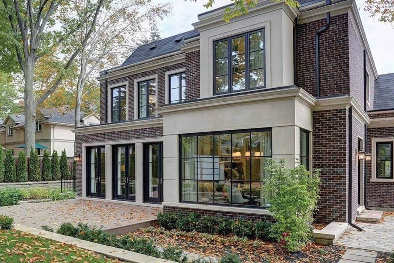 A contemporary two-story brick home with black-framed windows, beige trim, and a spacious patio area surrounded by trees and landscaping.