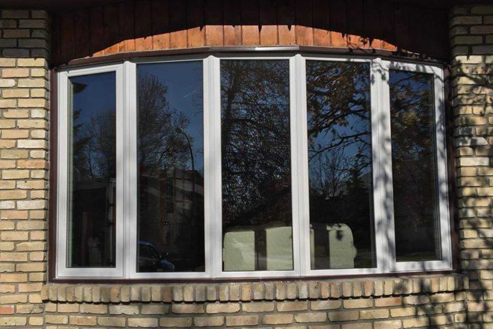 A white-framed curved bay window installed in a light brick wall, reflecting trees and sky on the glass surface.