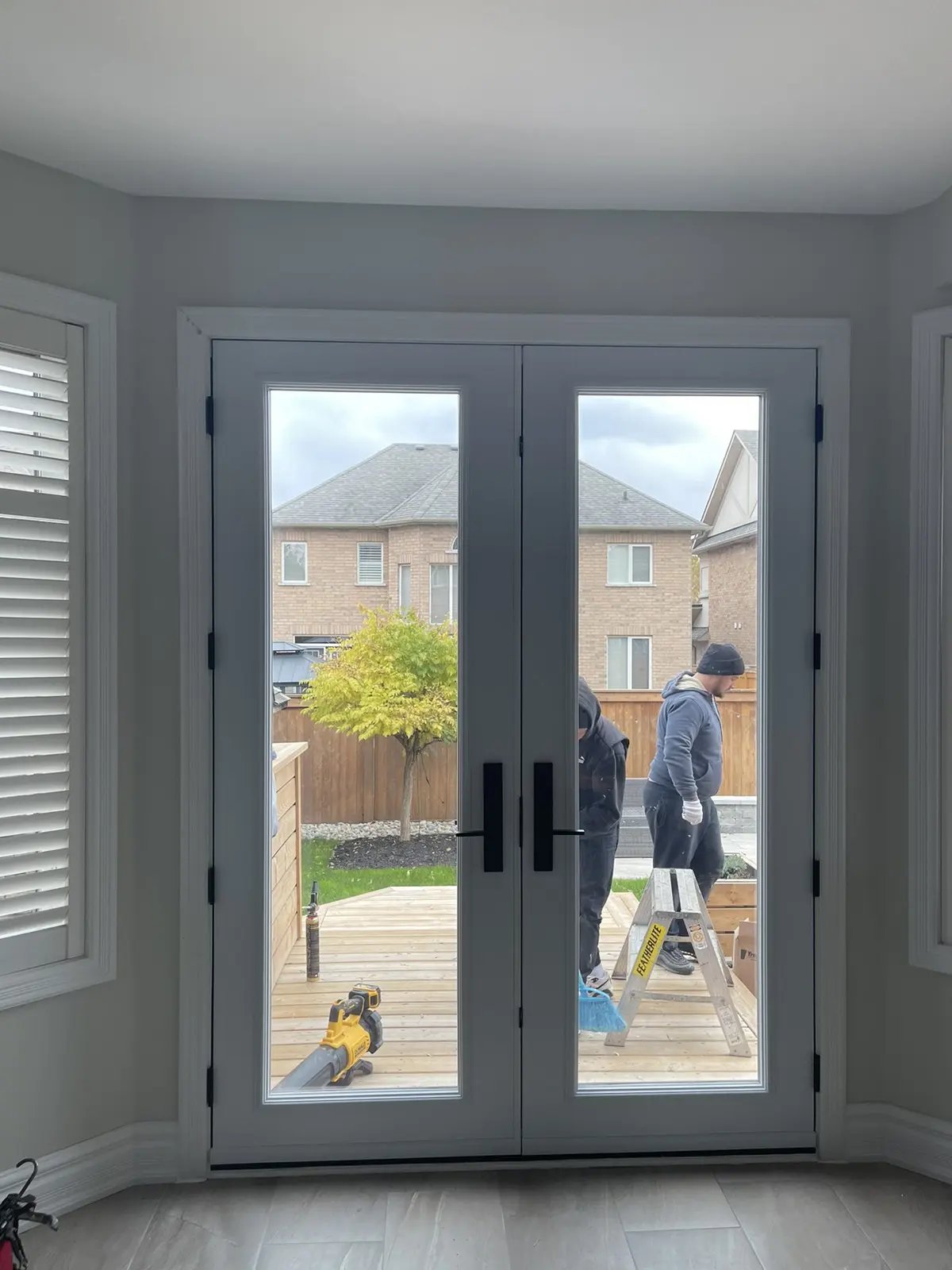 Interior view of modern white double patio doors with black handles, leading to a wooden backyard deck where two workers are standing near a ladder and tools. A small tree and neighboring brick houses are visible in the background.