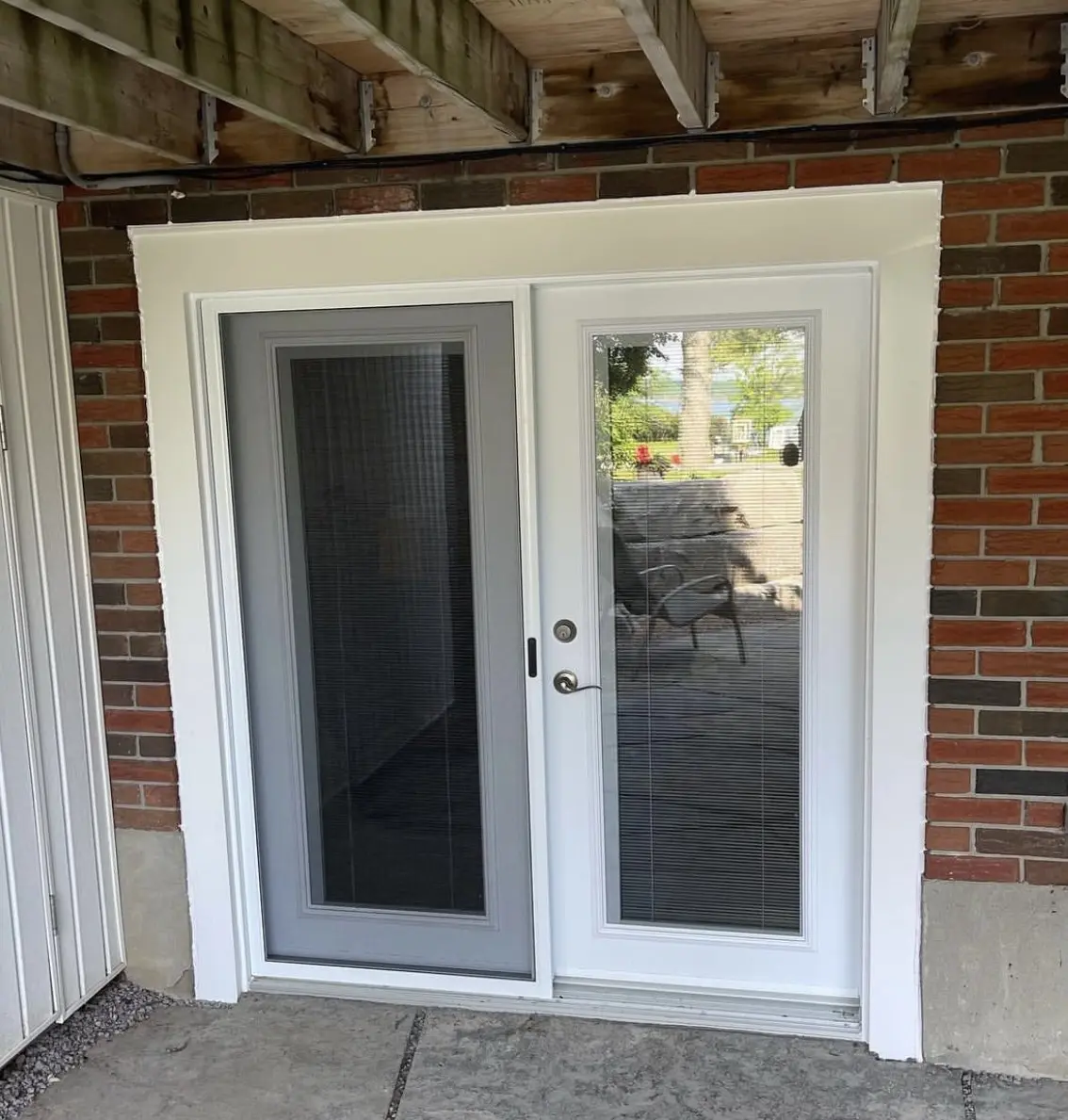 Exterior view of a white sliding patio door with built-in blinds between the glass panes, set in a red brick wall with a concrete base and wooden deck above.