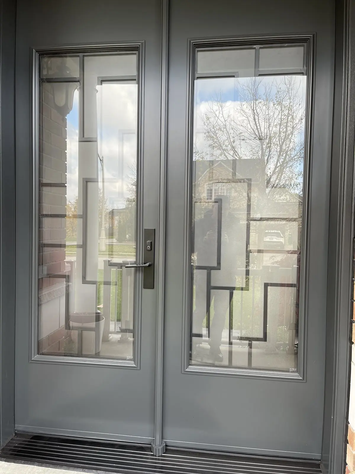 Modern gray double front doors featuring large vertical glass panels with a stylish geometric grid design, allowing a view of the front yard and street beyond.