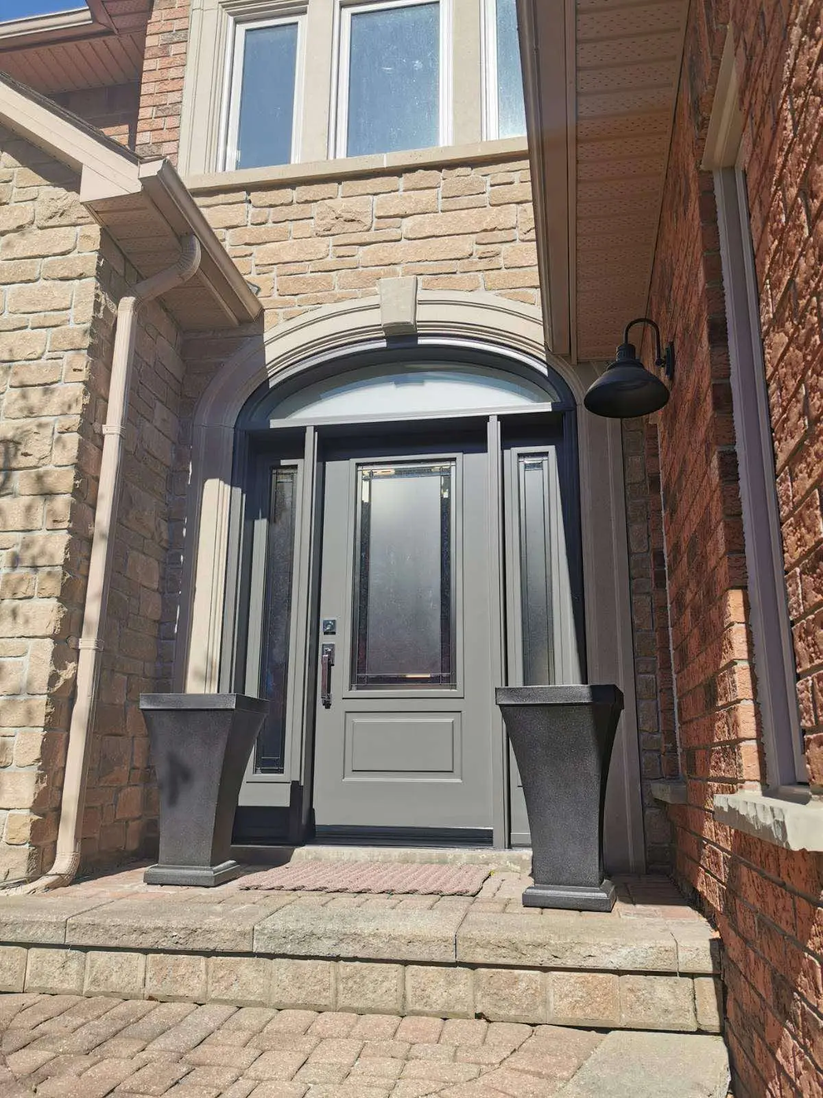 Grey front entry door featuring a central decorative glass panel, framed by two matching sidelites and topped with an arched transom window, set in a stone and brick façade with two large black planters on each side.