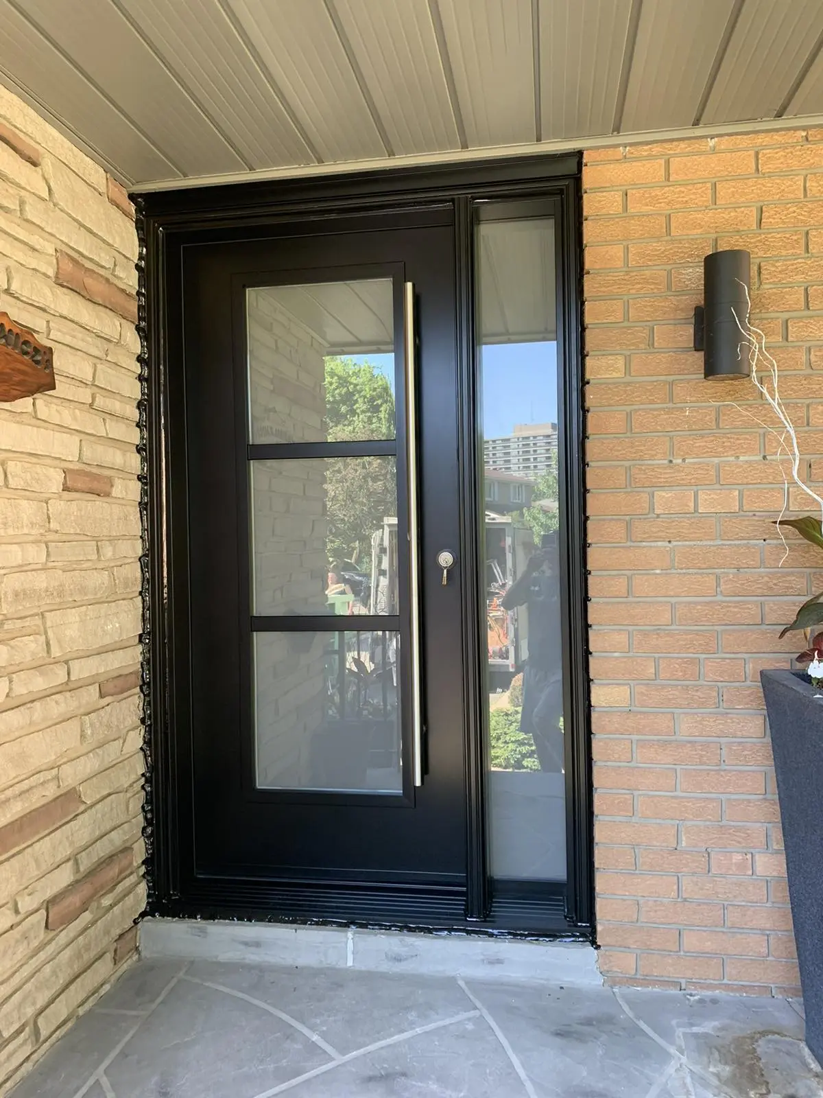 Sleek black front door with three horizontal frosted glass panels and a matching full-height side lite, installed between light tan brick and stone walls under a covered porch.