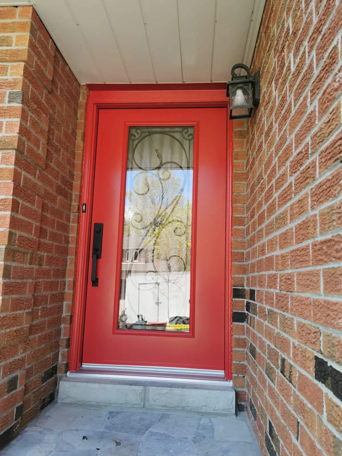Close-up of a bold red entry door featuring a central glass panel with elegant black wrought iron scrollwork, surrounded by red brick walls and topped with a black exterior light fixture.