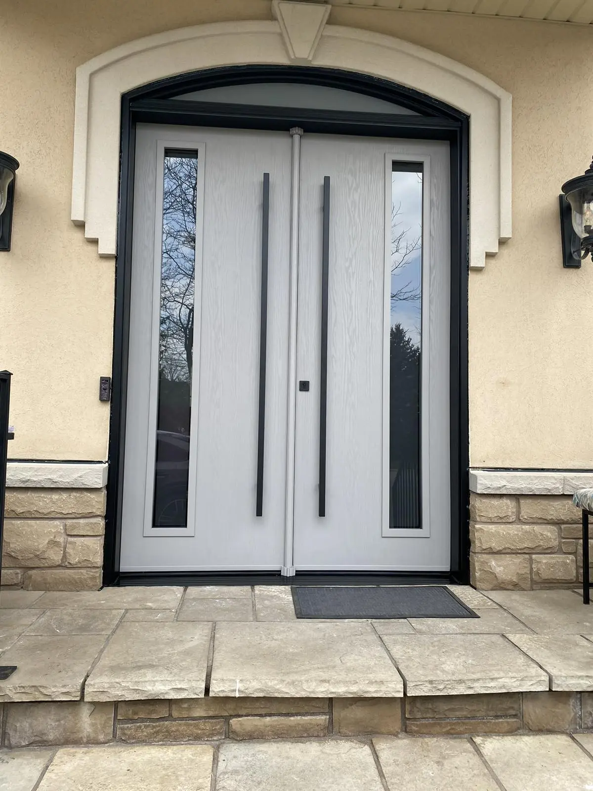 Front view of a modern light gray double door featuring narrow vertical glass inserts and sleek black handles, framed by stonework and a decorative arched transom window above.