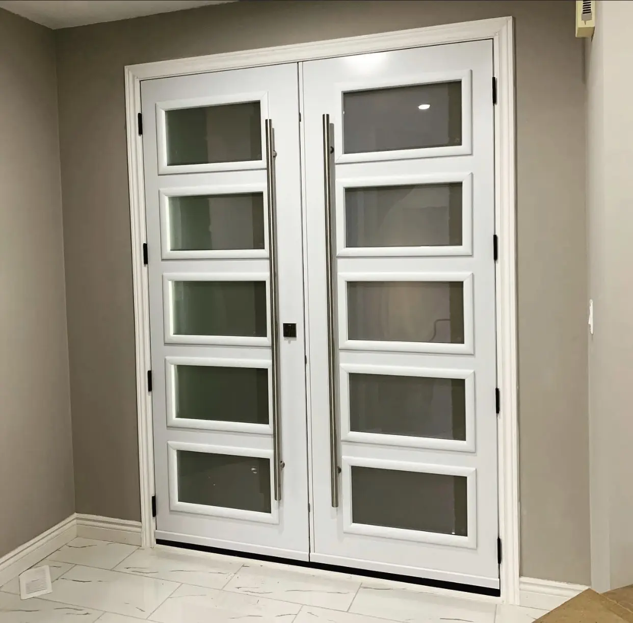 Contemporary white double front doors featuring vertical rectangular frosted glass panels in a symmetrical layout, set within a neutral-toned interior hallway.