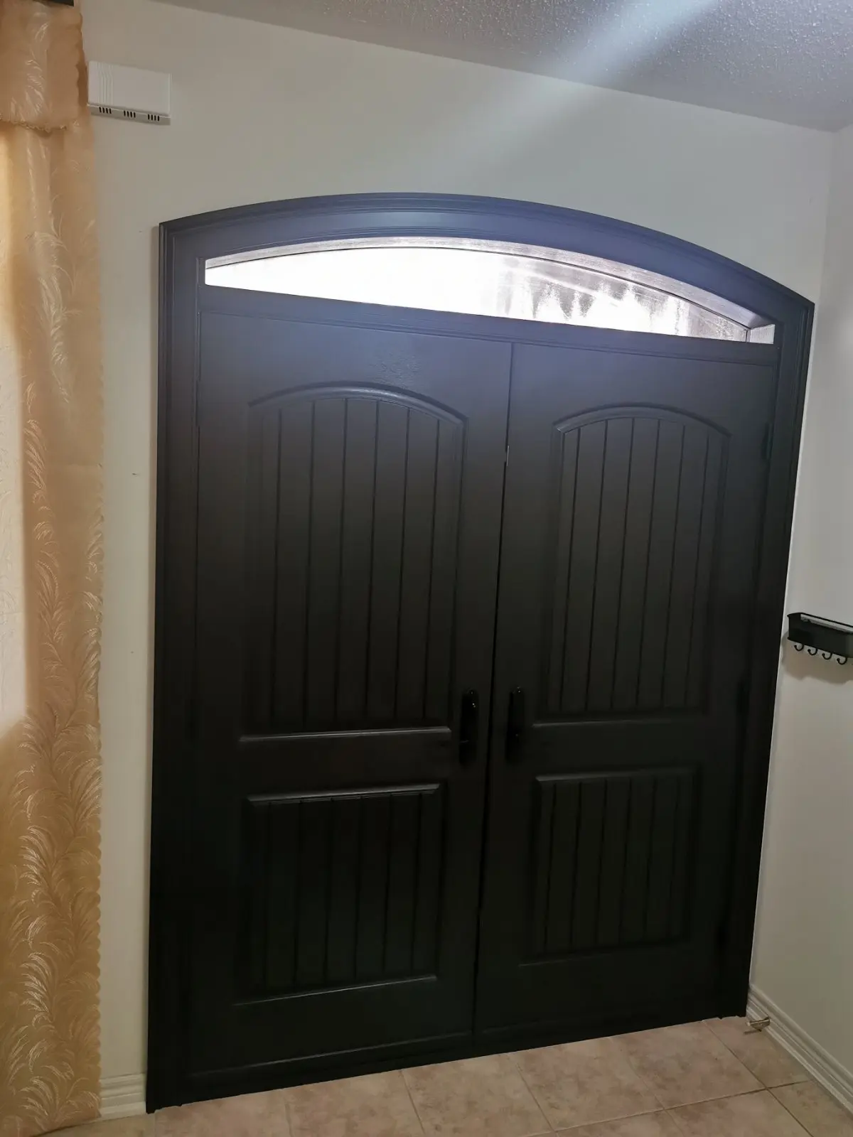 Interior view of a dark double front door with vertical panel detailing and a curved frosted glass transom window above, set against a white wall and beige tiled floor.
