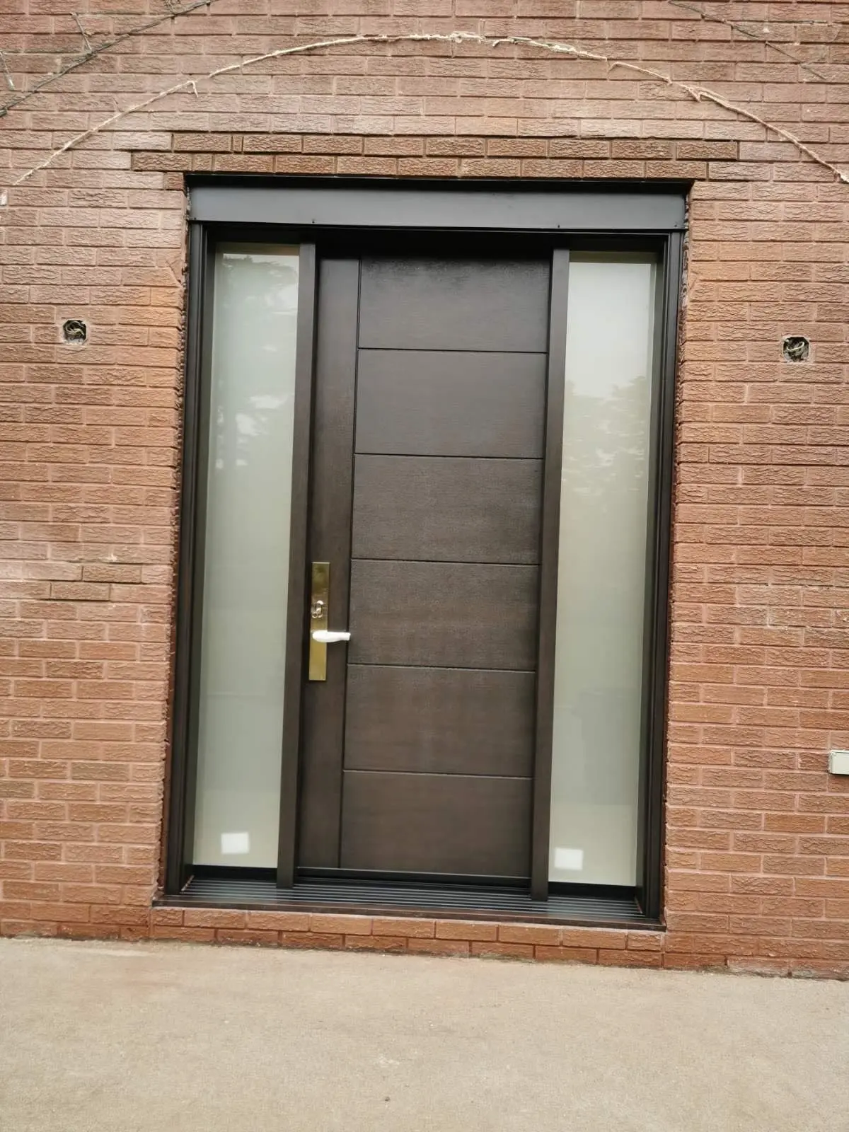 Sleek modern front entry door in dark brown wood finish, featuring horizontal panel design and frosted glass sidelights on both sides, set in a red brick exterior.