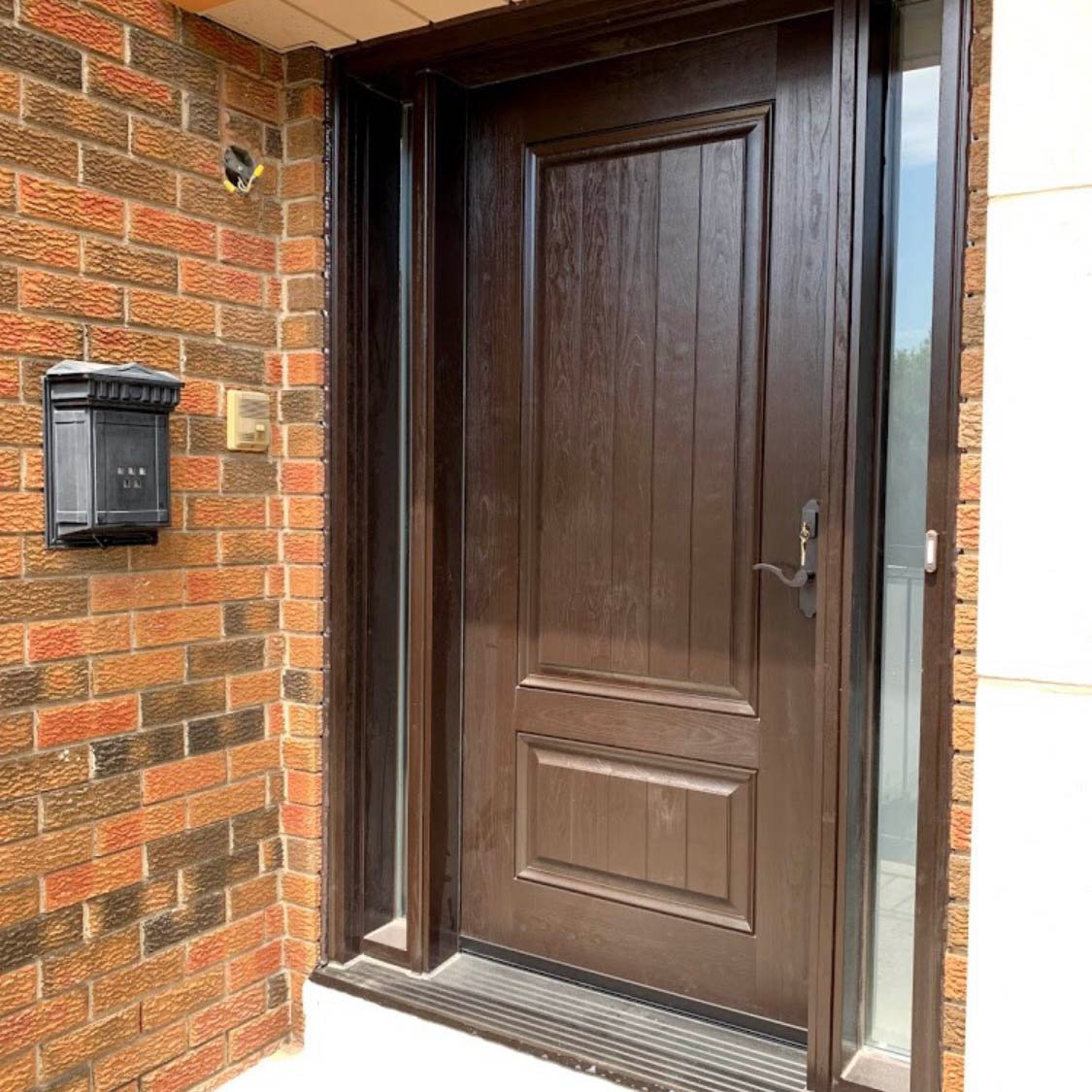 Modern dark wood front door featuring two sidelights and a black mailbox on a brick exterior wall.