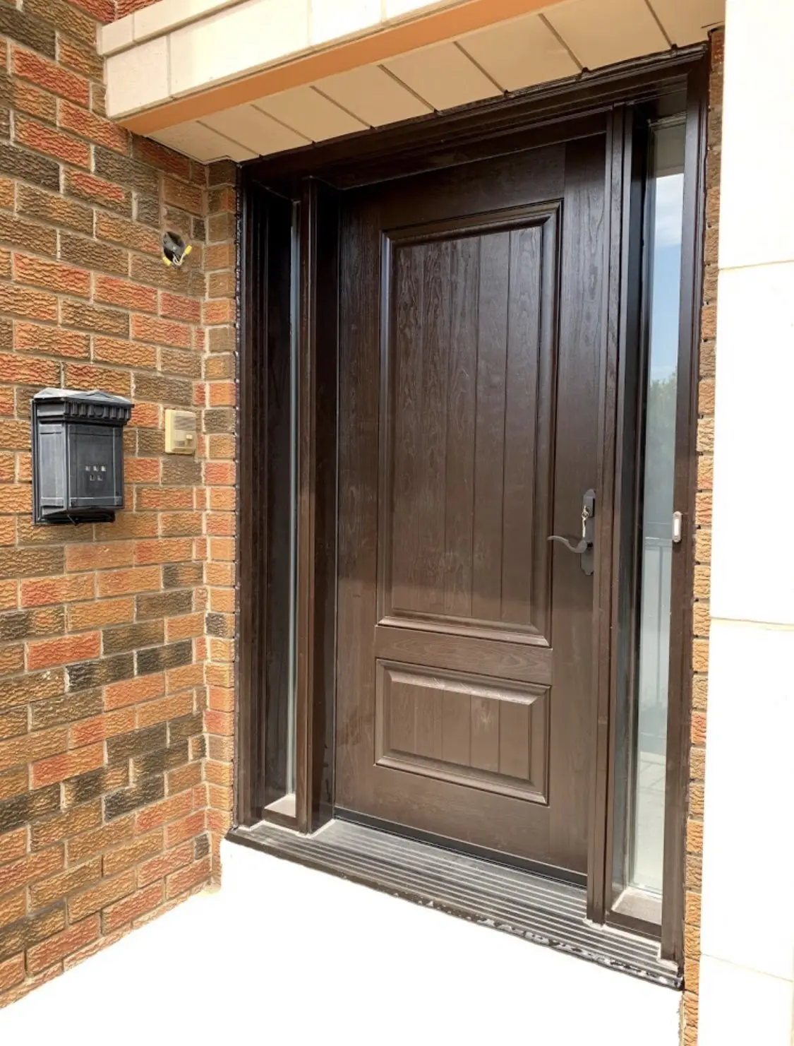 Front view of a modern dark brown wood-grain entry door with classic rectangular panel design, flanked by narrow glass sidelights on both sides. The door is set in a red brick exterior with a black mailbox and doorbell.
