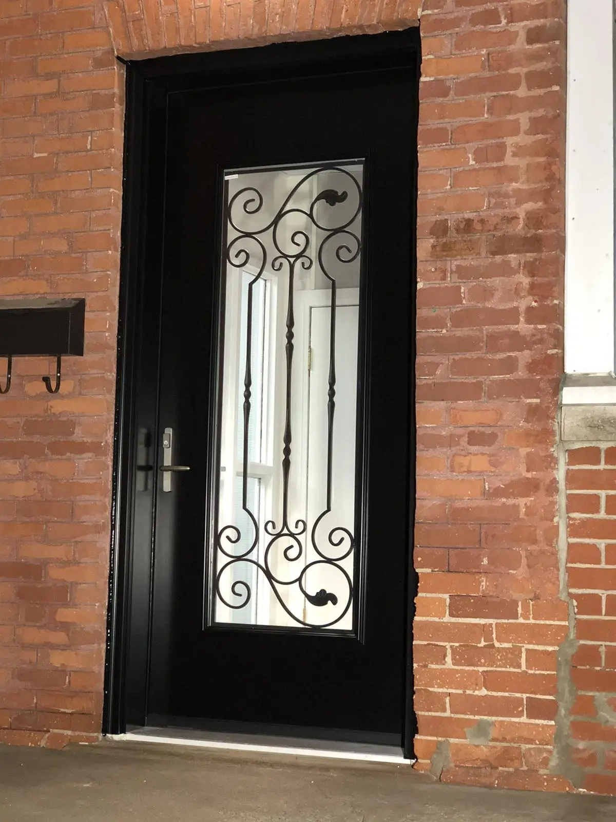 Black exterior entry door set in a red brick wall, featuring a tall glass panel with ornate black wrought iron scrollwork. The door has a modern silver handle and reflects an interior hallway with a white door.