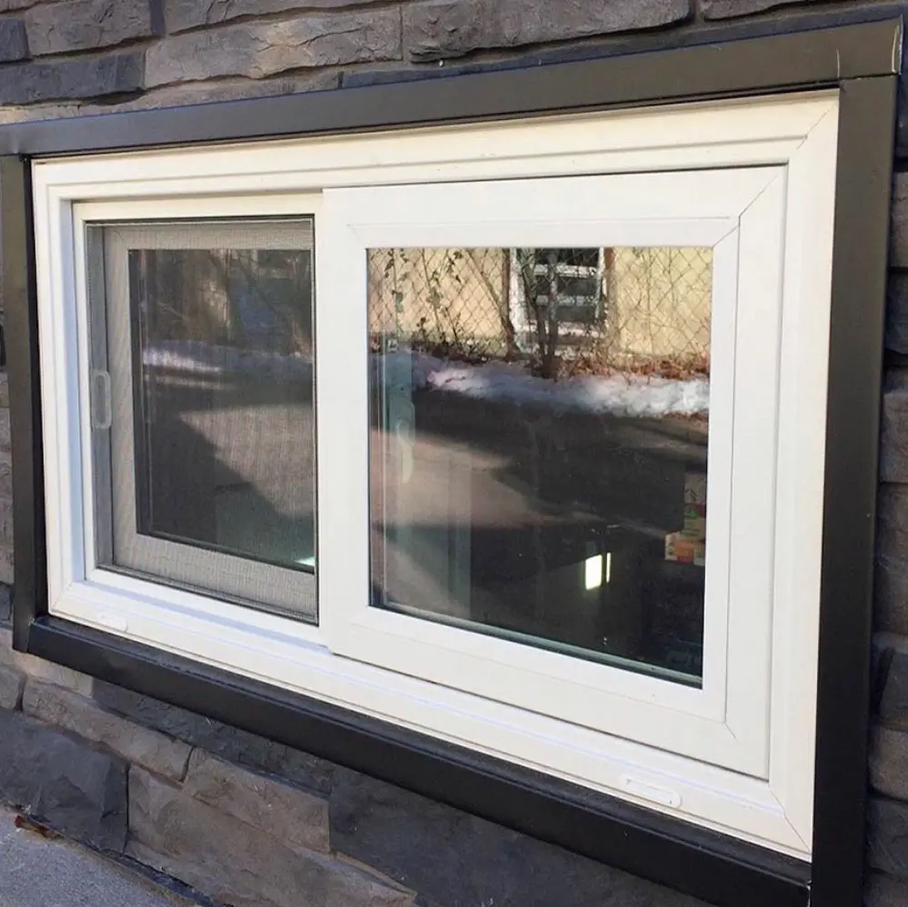 White-framed basement sliding window set in a dark stone exterior wall, with clear and screen panels reflecting a snowy backyard.