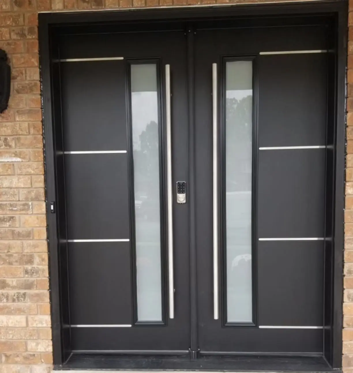 Modern double black front doors with vertical frosted glass inserts, stainless steel handles, and a digital lock keypad, set in a brick exterior wall.