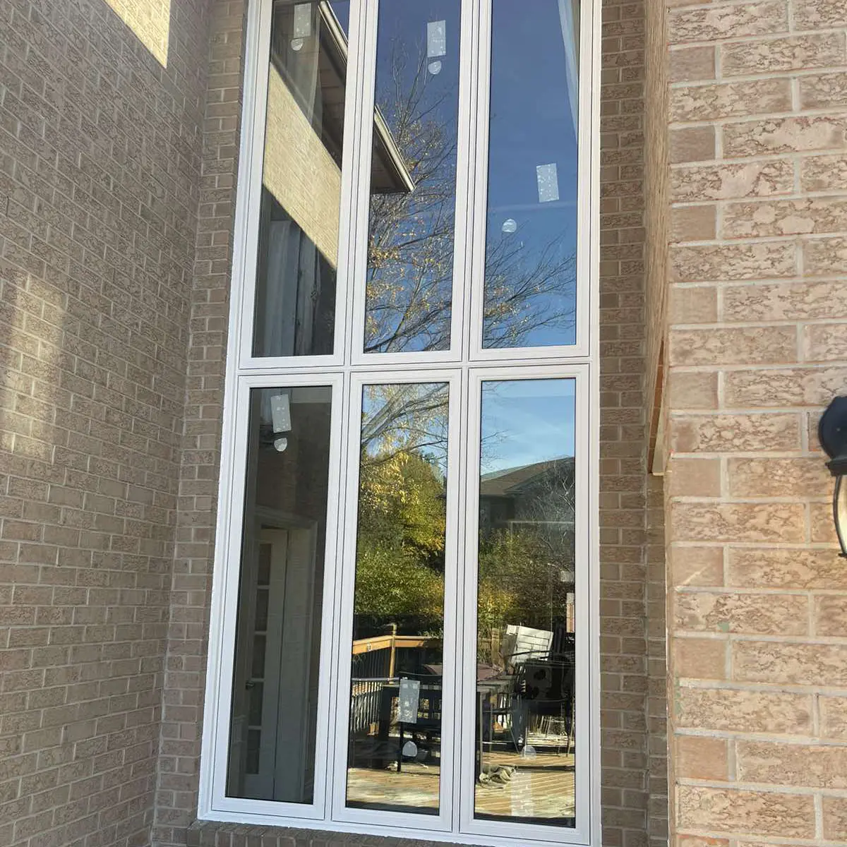 Tall multi-panel window with white frames on a brick exterior wall, reflecting trees, sky, and patio furniture.