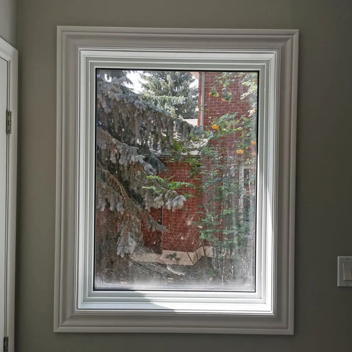 Interior view of a modern, white-trimmed window looking out to a red brick house and evergreen trees.