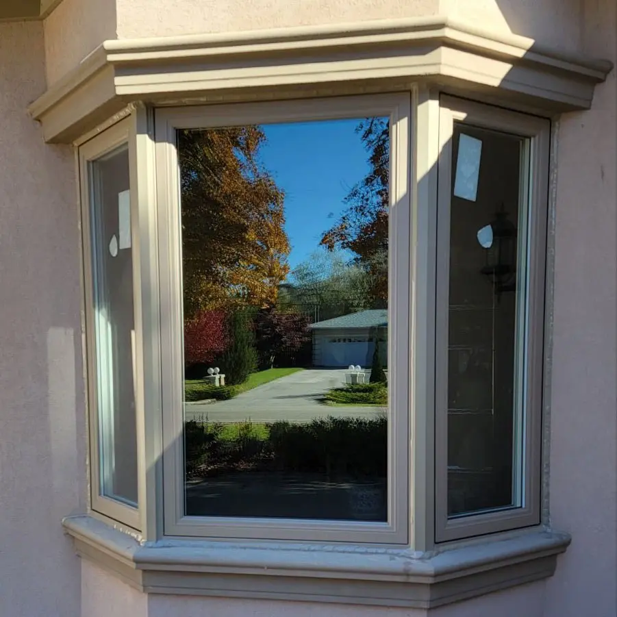 Three-panel bay window with beige frame reflecting a residential street and autumn trees.