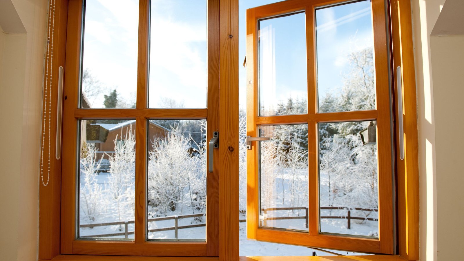 Open wooden-framed double windows revealing a snowy outdoor landscape with frosted trees, a wooden fence, and a house in the background under a clear blue sky.