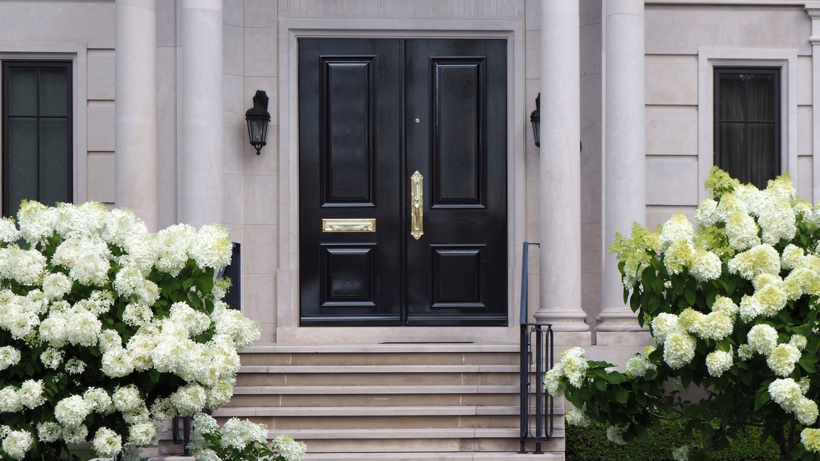 Elegant black double entry door with gold hardware, framed by white stone columns and blooming white hydrangeas.