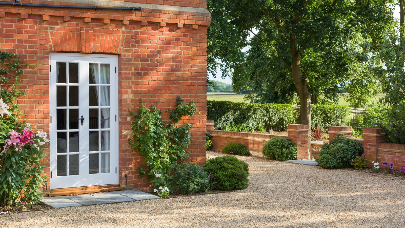 Exterior view of a traditional red brick house with newly installed white French doors opening to a garden with flowering plants, shrubs, and a gravel walkway.
