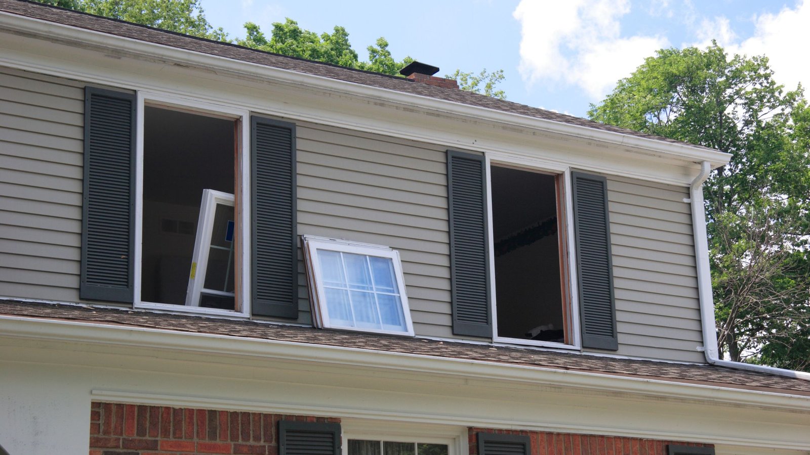 Second-story window replacement in progress on a residential home, with window frames removed and a new window resting on the roof edge.