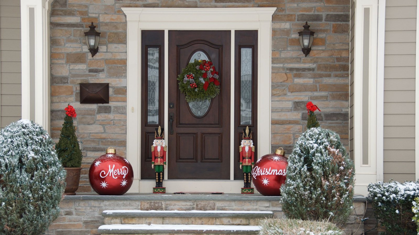 Front entrance of a home decorated for Christmas, featuring a dark wooden steel front door with a festive wreath, flanked by two nutcracker figures and large red ornaments reading "Merry Christmas."