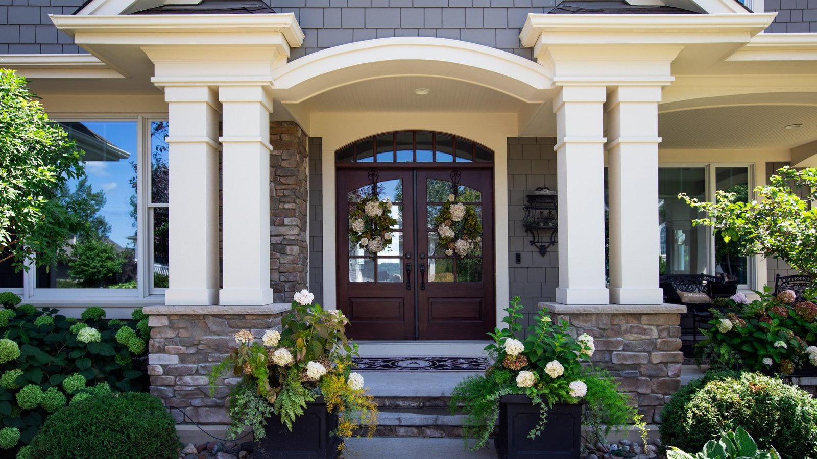 Elegant double front doors made of dark wood with decorative glass panels and floral wreaths, framed by stone columns and surrounded by lush greenery and symmetrical landscaping.
