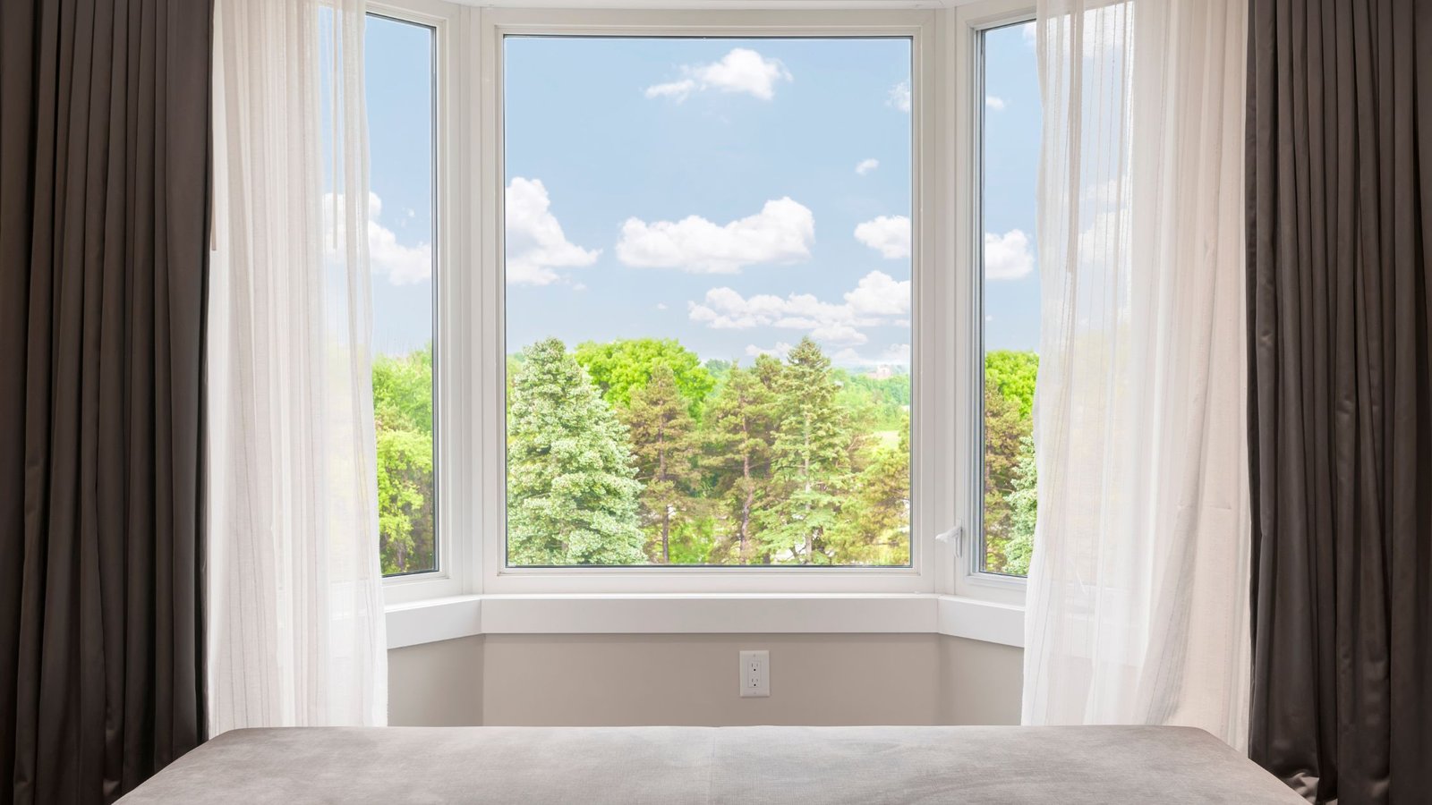 A large bay window with three glass panels framed by white curtains, offering a clear view of green trees and a bright, cloudy sky from inside a bedroom.