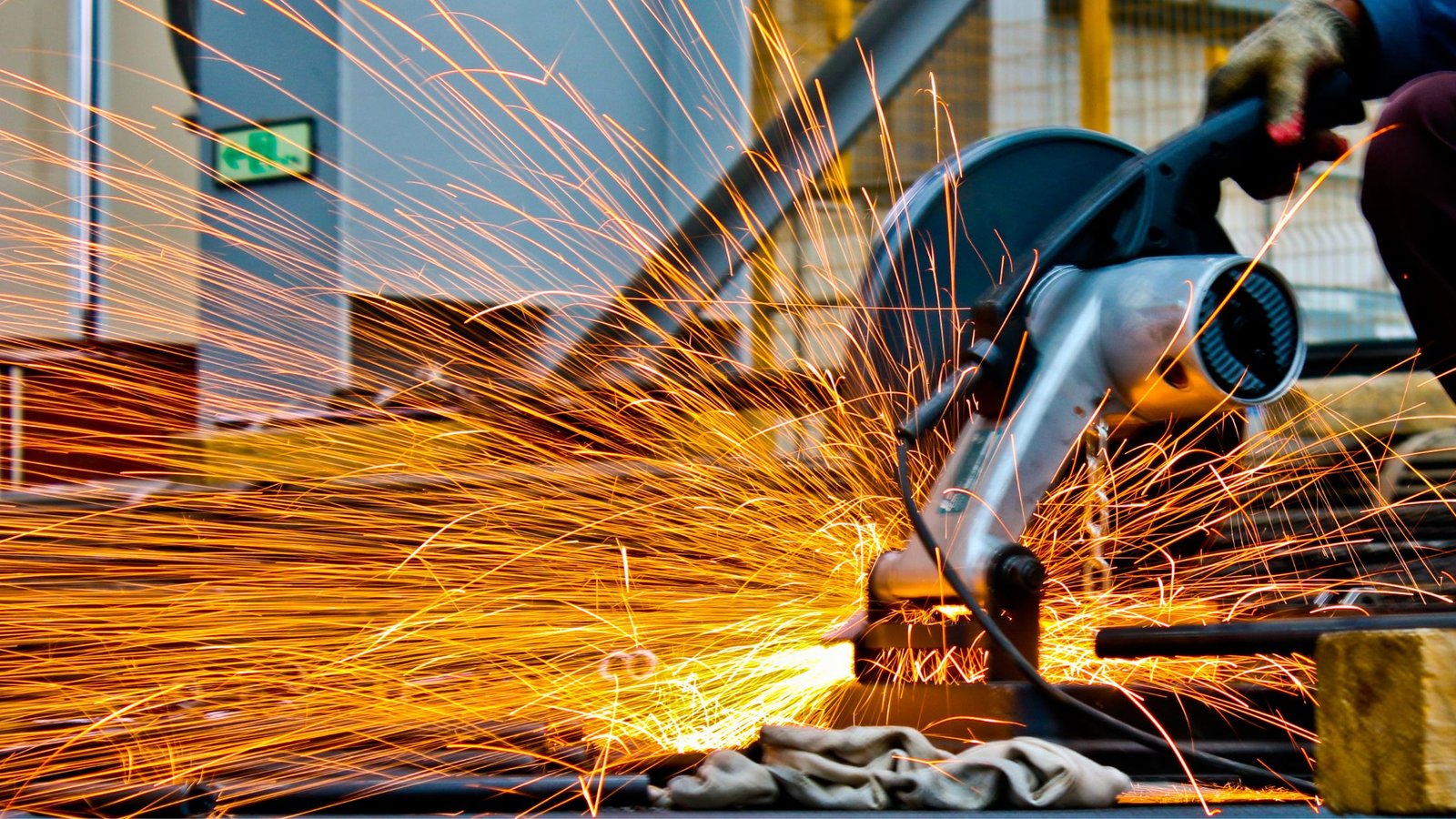 A close-up of a worker using a power metal-cutting saw, producing bright orange sparks while cutting through steel in an industrial setting.