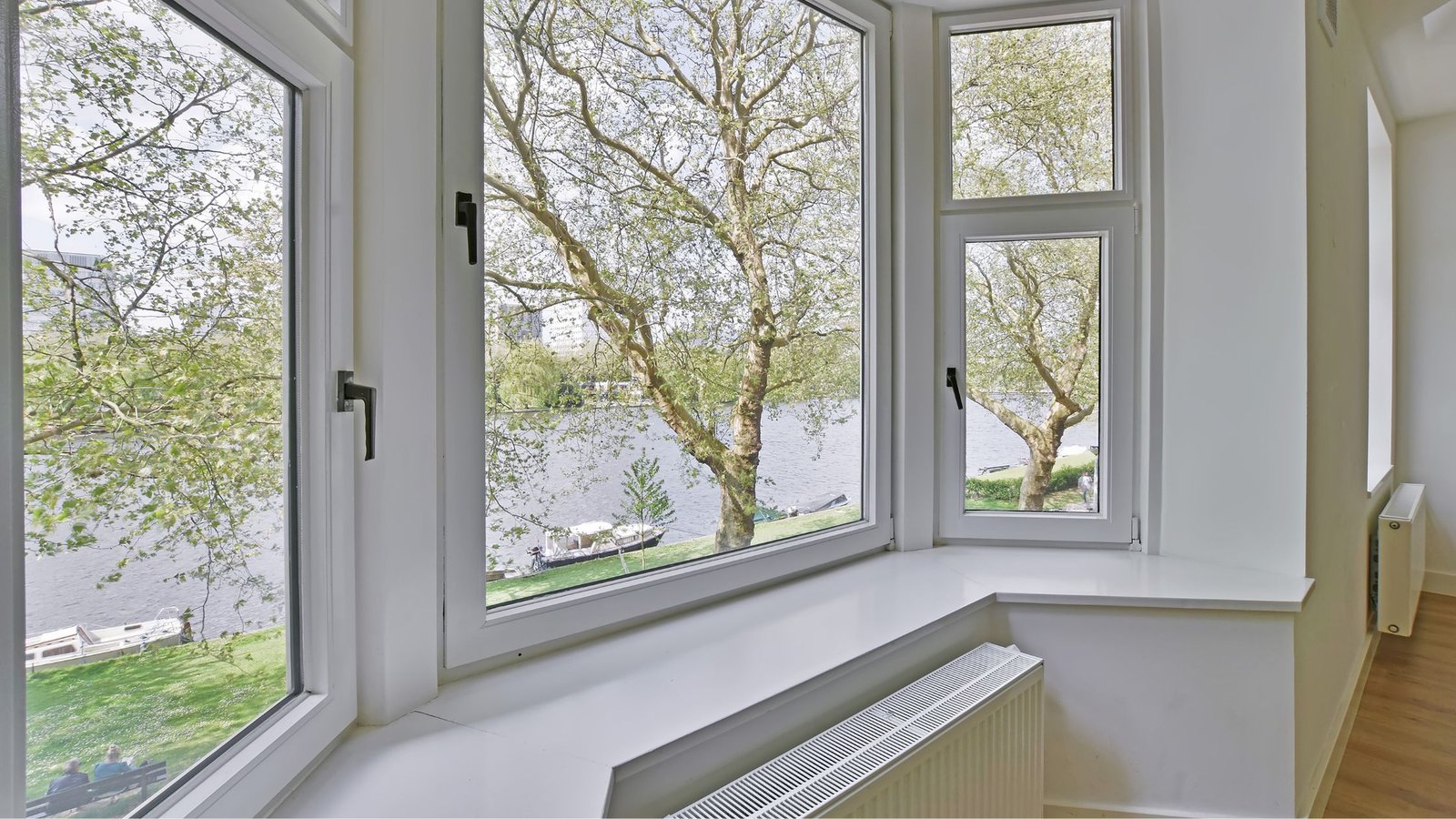Interior view of modern triple-pane windows with white frames, overlooking a river and green park with large leafy trees.