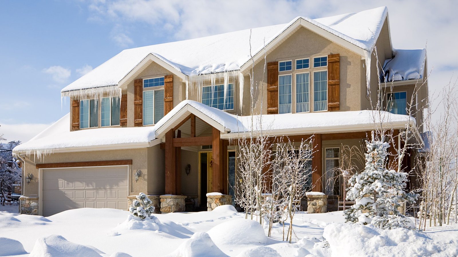 Modern two-story home in winter with snow-covered roof and large icicles hanging from eaves, illustrating how to stop ice buildup on your windows.